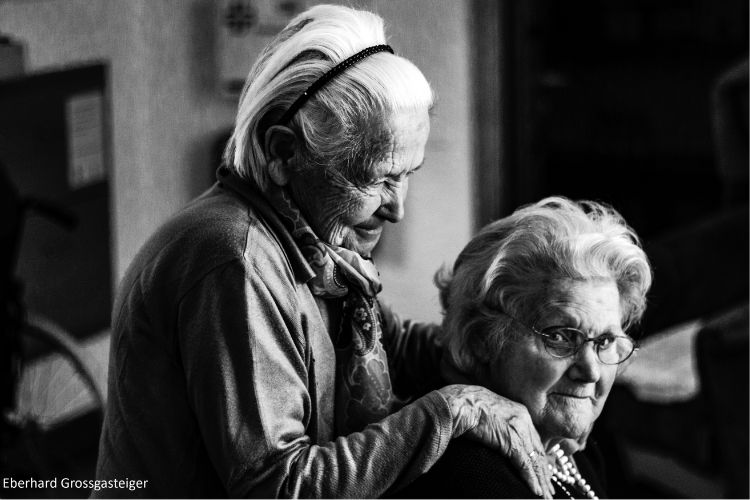 Two elderly women, one is sat on a chair looking at the camera and the other is stood behind her with her hands on her shoulder